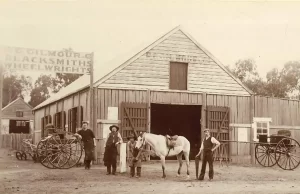 Historic black and white image of a blacksmith barn and four men in cowboy hats, a horse and various carts and equipment