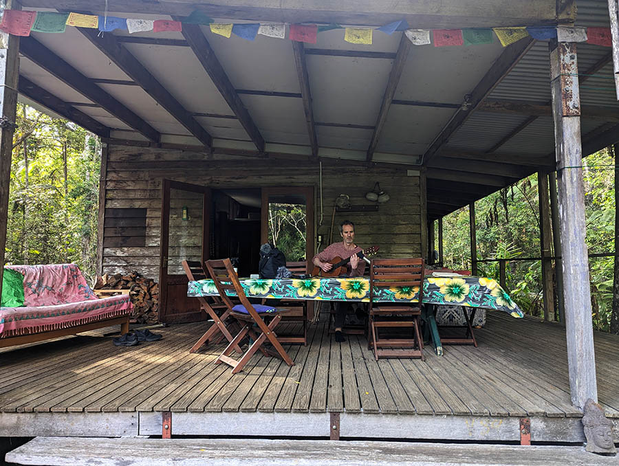 man playing guitar on the porch of a weatherbeaten little house in the rainforest.