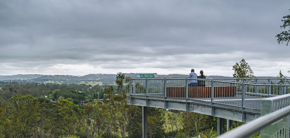 The Skywalk metal structure overlooking a rural landscape and a lot of sky. A couple is sitting and contemplating at the end of the platform.