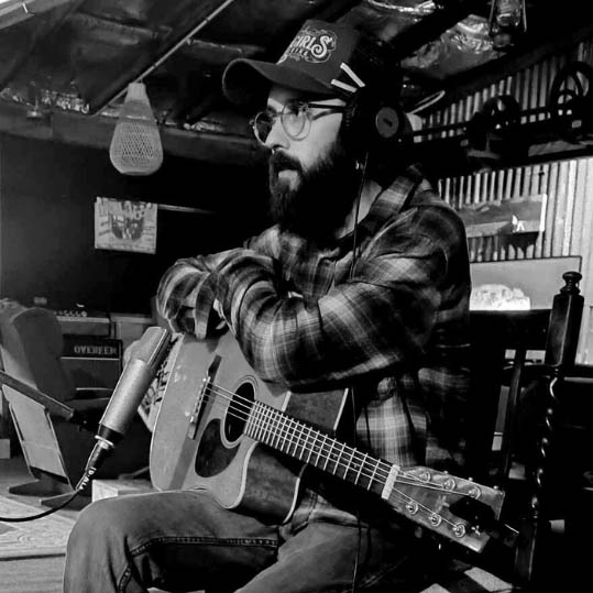 Black and white portrait of Michael Williams. He is in a recording environmentcap, beard, glasses a checked shirt and a guitar on his lap.