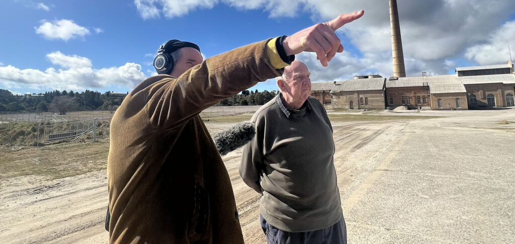 Man with headphones and microphone pointing into the distance with older man beside him